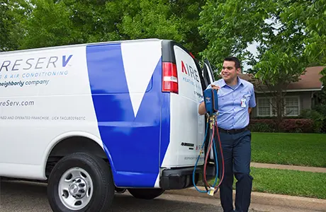 Male Aire Serv technician holding AC manifold gauge and hose set beside branded van on residential street.